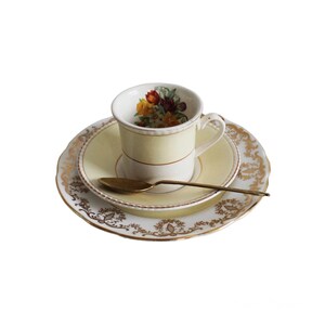 Puede incluir: Un juego de té vintage con taza, platillo y cuchara dorada. La taza y el platillo son de color amarillo pálido con ribetes dorados y diseños florales. El interior de la taza presenta un arreglo floral colorido. La cuchara es dorada.