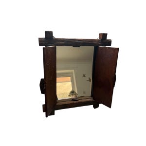 Op de afbeelding: Een rustieke spiegel met houten frame en twee scharnierende deuren. Het frame is gemaakt van donkerbruin, ruw bewerkt hout, met een rechthoekige spiegel in het midden. De deuren staan open en tonen de weerspiegeling van een kamer. De spiegel is ontworpen om aan de muur te worden bevestigd.