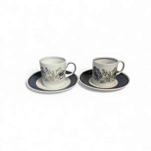 May include: Two white ceramic coffee cups with matching saucers. Each cup and saucer set features a floral design in shades of blue and green. The saucers have a dark blue border. The cups have a silver handle.