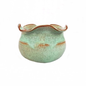 May include: A small, round ceramic bowl with a ruffled rim. The bowl is glazed in a mottled turquoise color with brown accents around the rim and mid-section. The handmade pottery has a rustic, organic aesthetic.