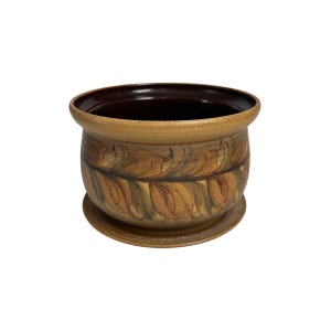 May include: A round ceramic planter with a brown interior and a light brown exterior. The exterior features a repeating pattern of stylized leaves in shades of brown and green. The planter sits on a matching saucer.