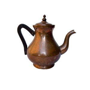 May include: A copper teapot with a dark handle and lid. The teapot has a rounded body and a curved spout. The handle is made of a dark material, possibly wood or a composite. The lid is topped with a small, decorative finial.