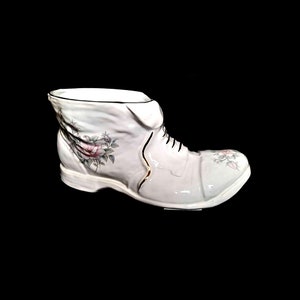 Puede incluir: Un jarrón blanco de cerámica con forma de bota y acabado brillante. La bota presenta cordones negros, un adorno dorado y diseños florales con rosas rosas y grises. El jarrón está sobre un fondo negro.