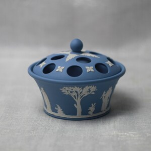 May include: A blue ceramic flower frog with a lid. The lid has several circular holes and is adorned with white floral and leaf designs. The base features white relief images of figures and trees. The overall design is a classic, elegant style.