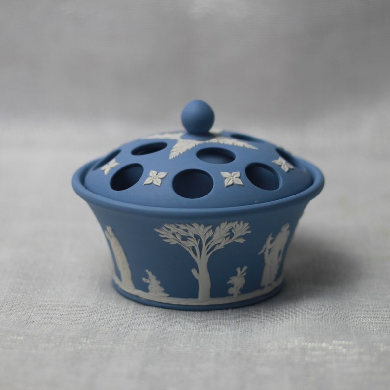 May include: A blue ceramic flower frog with a lid. The lid has several circular holes and is adorned with white floral and leaf designs. The base features white relief images of figures and trees. The overall design is a classic, elegant style.