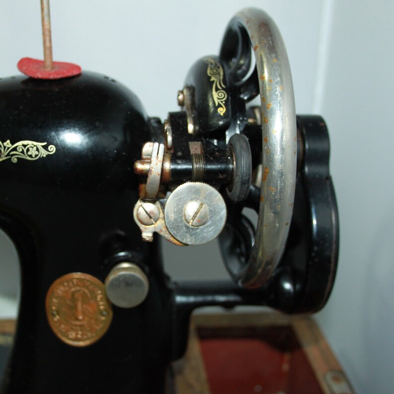 May include: Close-up of a vintage black Singer sewing machine. The machine features a large hand wheel, decorative gold accents, and a round metal plate with text. The machine's components show signs of age and use.