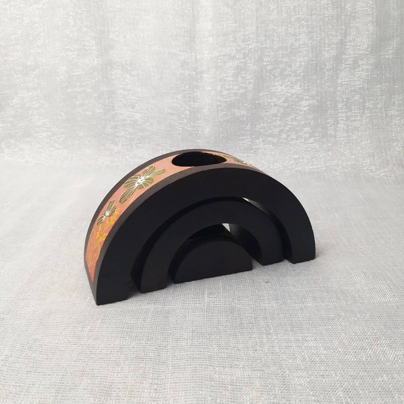 K&ouml;nnte beinhalten: Schwarzes Holzdekor in Regenbogenform mit floralem Design in Rosa, Gelb und Gr&uuml;n. Der Artikel hat oben ein Loch und besteht aus drei gestapelten B&ouml;gen.