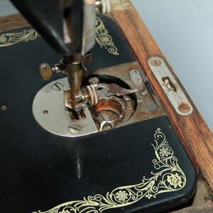 May include: Close-up of an antique black sewing machine with gold floral accents. The needle and bobbin area are visible, showing signs of age and use. The wooden base and metal components have a vintage aesthetic.