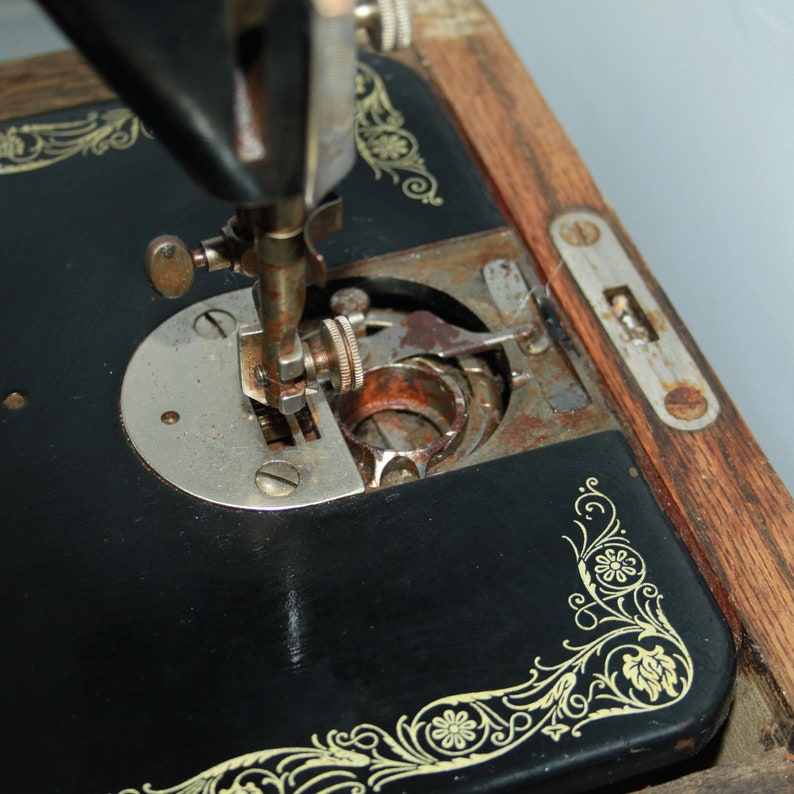 May include: Close-up of an antique black sewing machine with gold floral accents. The needle and bobbin area are visible, showing signs of age and use. The wooden base and metal components have a vintage aesthetic.