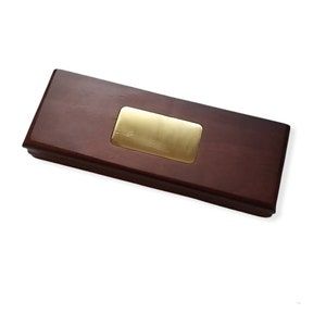 May include: A rectangular wooden box with a dark brown finish and a gold-colored metal plate in the center. The box has a simple, elegant design, suitable for various uses.