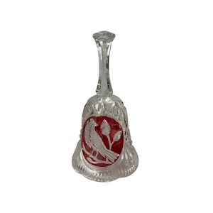 May include: A clear crystal bell with a long handle and a flared base. The bell features a red panel with an etched design of a bird perched on a branch. The bell has a faceted handle and a scalloped edge.