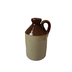 Könnte beinhalten: Kleiner, antiker Steinzeugkrug mit Henkel. Der Krug ist zweifarbig, mit einem hellbraunen Sockel und einem dunkelbraunen Oberteil und Henkel. Der Krug hat einen runden Körper und einen kleinen Ausguss.