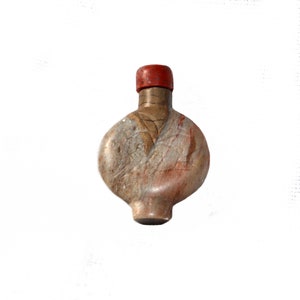 Puede incluir: Botella pequeña de estilo antiguo, con cuerpo redondeado y tapón rojo. La botella tiene un esquema de color moteado y terroso con tonos de beige, marrón y rojo. El diseño sugiere una estética vintage o artesanal, adecuada para exhibición o colección.
