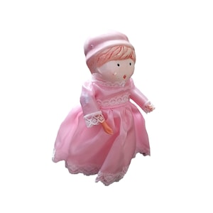 May include: A vintage porcelain doll in a pink dress with white lace trim. The doll has a matching pink bonnet and painted facial features. The dress has long sleeves and a full skirt. The doll's arms are visible.
