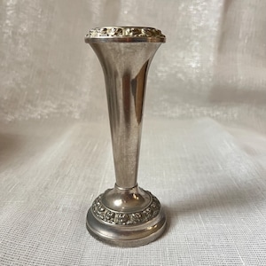 May include: A small, silver-colored bud vase with a flared top and a detailed base. The vase has a slender, elongated body and a decorative rim. The base features an embossed floral design. The vase is approximately 6 inches tall.
