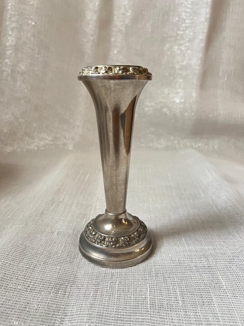 May include: A small, silver-colored bud vase with a flared top and a detailed base. The vase has a slender, elongated body and a decorative rim. The base features an embossed floral design. The vase is approximately 6 inches tall.