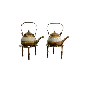 May include: Two vintage brass teapots with white and gold detailing. Each teapot features a curved spout, a handle, and rests on a small brass stand with legs. The teapots are set against a white backdrop.