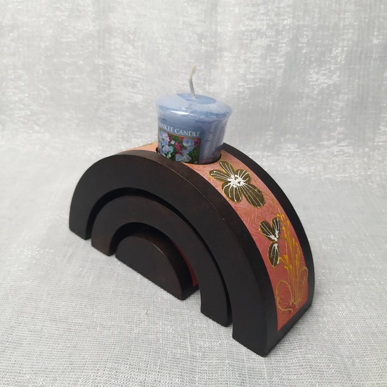 K&ouml;nnte beinhalten: Ein dunkelbrauner Kerzenhalter aus Holz in Regenbogenbogen-Design. Der Halter hat ein Blumenmuster in Rosa und Gold. Eine hellblaue Yankee Candle ist oben platziert.