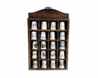 May include: A dark wooden display case filled with sixteen decorative thimbles. Each thimble is white with a blue or black illustration of a landmark or scene. The case has a curved top and a small clasp.