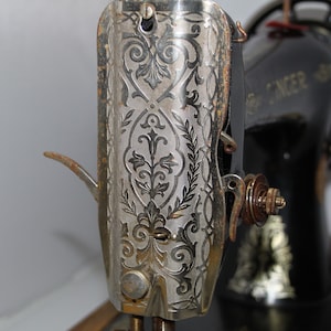 May include: Close-up of an antique Singer sewing machine. The silver-colored metal casing features an ornate floral design. The machine's black body is visible in the background, with the Singer logo. The machine shows signs of age and use.