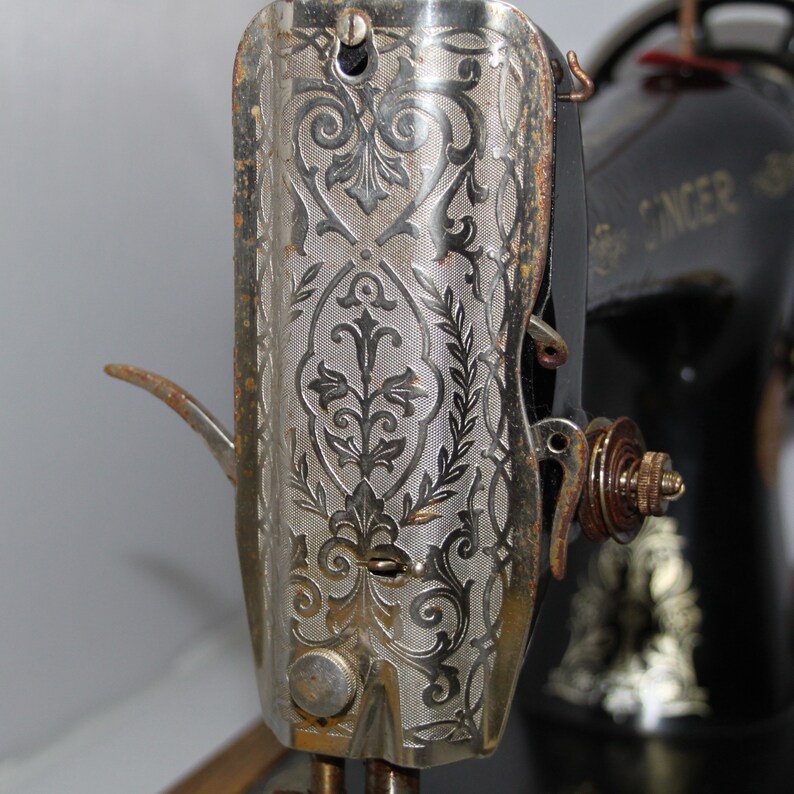 May include: Close-up of an antique Singer sewing machine. The silver-colored metal casing features an ornate floral design. The machine's black body is visible in the background, with the Singer logo. The machine shows signs of age and use.