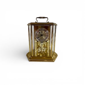 Può includere: Un orologio rettangolare color oro con una maniglia sulla parte superiore. Il quadrante dell'orologio ha numeri romani e un pendolo decorativo. I lati sono in vetro con un motivo bianco. L'orologio poggia su una base dorata.