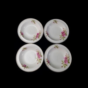 May include: Four white ceramic bowls with a pink rose and green leaf design. Each bowl has a beaded gold rim. The bowls are arranged in a square on a black background.