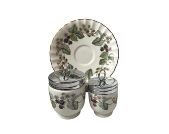 Könnte beinhalten: Ein Vintage-Eierbecher-Set mit zwei Eierbechern mit silbernen Deckeln und einer passenden Untertasse. Das Set ist mit einem botanischen Design aus grünen Blättern und Brombeeren auf cremefarbenem Hintergrund verziert. Die Untertasse hat einen Goldrand.