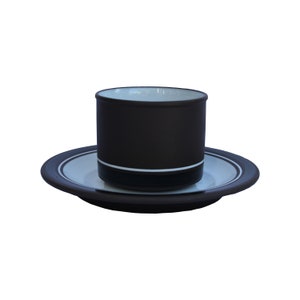 Könnte beinhalten: Ein dunkelbraunes Kaffeebecher- und Untertassen-Set. Der Becher ist zylindrisch mit weißem Innenraum und einem weißen und schwarzen Band am Boden. Die Untertasse hat ein passendes Design mit weißer Mitte und braunem Rand.