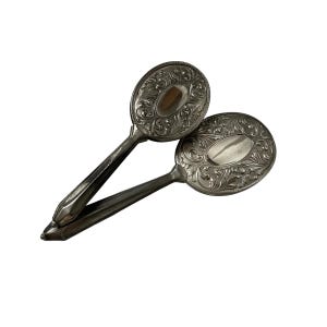 May include: Two antique silver-toned hand mirrors. Each mirror has an oval-shaped head with an ornate, floral design. The handles are long and slender, with a pointed end. The mirrors are displayed against a white background.