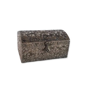 May include: A small, silver-coloured chest with a curved lid. The surface is intricately decorated with a floral pattern. A decorative clasp secures the front. The chest is likely made of metal and is designed for storing small items.