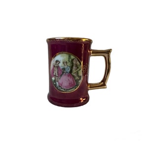 May include: A burgundy ceramic mug with a gold handle and rim. The mug features a painted oval scene of a couple in period clothing. The handle has a unique, ornate design. The mug is likely vintage or antique.