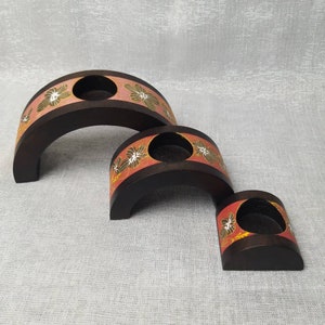 K&ouml;nnte beinhalten: Drei unterschiedlich gro&szlig;e Kerzenhalter aus Holz. Jeder hat eine dunkelbraune, gew&ouml;lbte Basis mit einer kreisf&ouml;rmigen &Ouml;ffnung f&uuml;r eine Kerze. Die Oberseite jedes Halters ist mit einem Band aus orangefarbenen und wei&szlig;en Blumenmustern verziert.