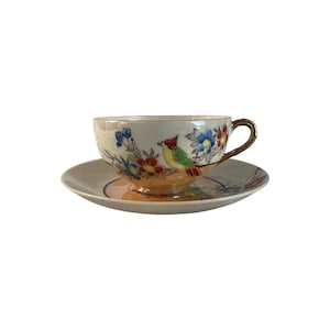 Op de afbeelding: Een vintage theekop en schotel set. De kop en schotel zijn wit met een geschilderd ontwerp van een vogel, bloemen en gouden rand. De vogel is geel, groen en rood. De bloemen zijn blauw, rood en geel.