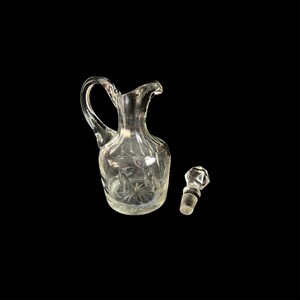 May include: A clear glass cruet with a handle and a decorative stopper. The cruet has a starburst design on the bottom and etched floral patterns. The stopper is faceted. The cruet is approximately 6 inches tall.