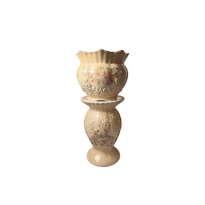 May include: A cream-coloured ceramic planter with a scalloped rim sits atop a matching pedestal base. Both pieces feature embossed scrollwork patterns and a central floral decal with pink and green tones, finished with a subtle gold-toned trim.