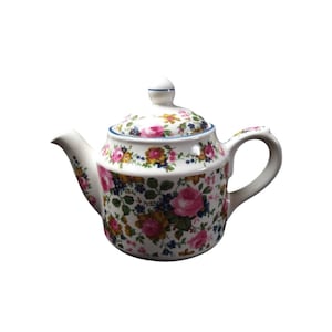 Könnte beinhalten: Weiße Keramik-Teekanne mit floralem Muster aus rosa Rosen, gelben und blauen Blumen und grünen Blättern. Die Teekanne hat einen gebogenen Ausguss, einen Henkel und einen Deckel mit einem kleinen Knauf. Ein blauer Rand akzentuiert den Deckel und den Boden.