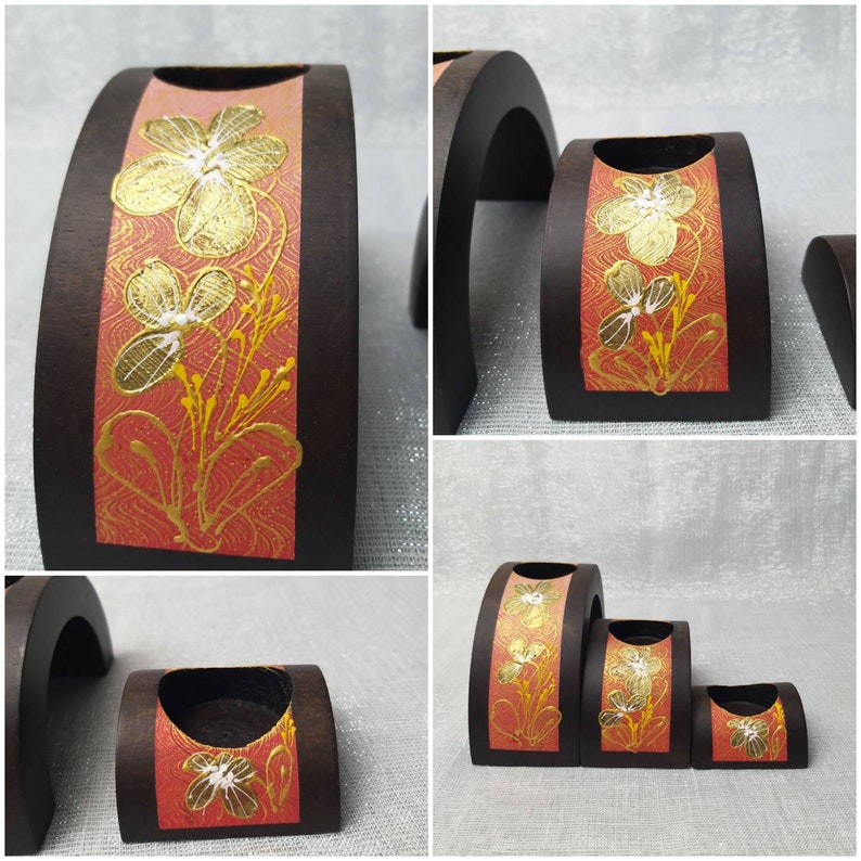 K&ouml;nnte beinhalten: Set aus drei h&ouml;lzernen Kerzenhaltern. Jeder Halter hat eine dunkelbraune, halbkreisf&ouml;rmige Form mit einem roten und goldenen Blumendesign auf der Vorderseite. Das florale Design zeigt goldfarbene Blumen und Stiele. Die Kerzenhalter sind unterschiedlich gro&szlig;.