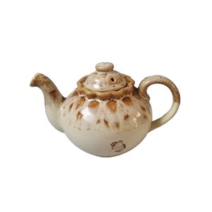 May include: A cream-coloured ceramic teapot with a rounded body and a lid. The teapot has a brown speckled pattern and a curved handle. The lid has a small knob and a decorative rim. A small label is visible on the bottom.