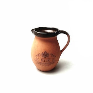 May include: A terracotta ceramic milk jug with a dark brown rim and handle. The jug features a decorative emblem with the word "MILK" and "Royal Barum Ware" printed on the front. The jug has a classic, rounded shape, suitable for serving milk.