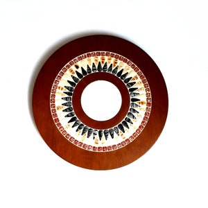 May include: A round, wooden decorative piece with a central hole. The outer edge is a dark brown, while the inner ring features a mosaic-like pattern of black, cream, and reddish-brown shapes. The design is symmetrical and geometric.