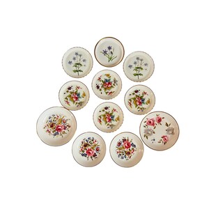 May include: A collection of small, white ceramic plates with floral designs. The plates feature various colorful flower arrangements, including roses and other blooms, with gold rims. Some plates have single flower sprigs.