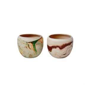 May include: Two ceramic bowls with a marbled design. One bowl features green, orange, and cream swirls, while the other has brown and cream patterns. Both have a rounded shape and a light brown interior.