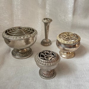 May include: Four silver flower arranging vases and holders. The collection includes a tall vase, two bowl-shaped holders, and a small, round holder. Each piece has a decorative, floral-inspired design and a silver finish.