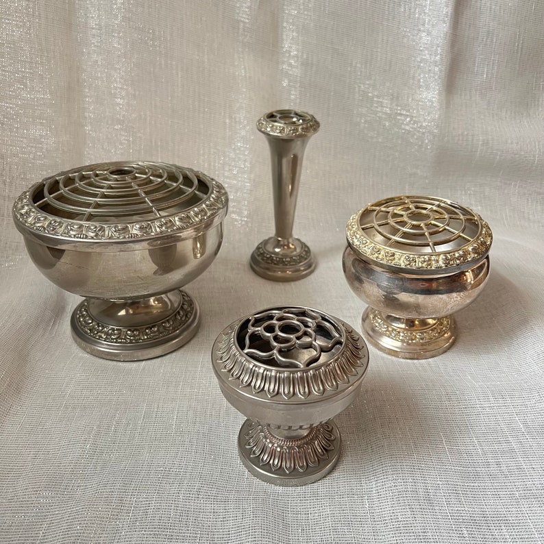 May include: Four silver flower arranging vases and holders. The collection includes a tall vase, two bowl-shaped holders, and a small, round holder. Each piece has a decorative, floral-inspired design and a silver finish.