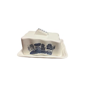 May include: White ceramic butter dish with a blue and white Willow pattern design. The dish has a square shape with a lid and a handle. The base is rectangular with a slightly raised edge.