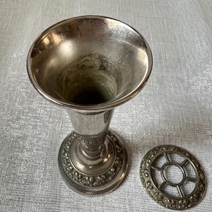 May include: A silver-toned vase with a flared top and a decorative base. A matching silver-toned floral frog sits beside the vase. The vase and frog have an aged appearance.