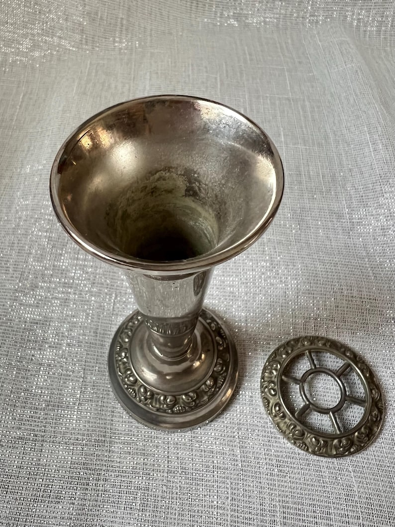 May include: A silver-toned vase with a flared top and a decorative base. A matching silver-toned floral frog sits beside the vase. The vase and frog have an aged appearance.