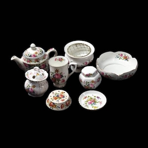 May include: A collection of white porcelain tableware with a floral rose pattern. The set includes a teapot, mug, sugar bowl, small jar, bowl, and a small plate. The edges of the bowl and plate have a gold trim.