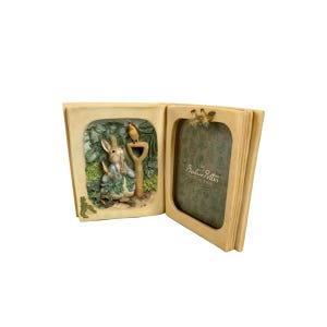 Puede incluir: Un marco de fotos en forma de libro abierto de color crema. El lado izquierdo presenta un relieve 3D de un conejo con una pala y un pájaro. El lado derecho tiene una abertura para fotos con el texto "The Beatrix Potter Collection".